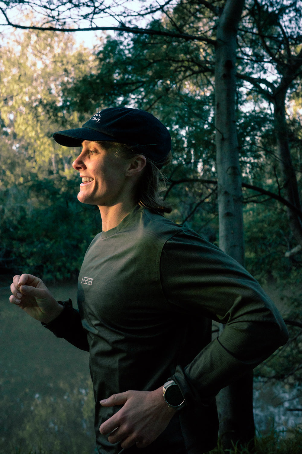 Runner in the Long Note LS Performance Tee in Olive and a black cap, side view on a riverside trail