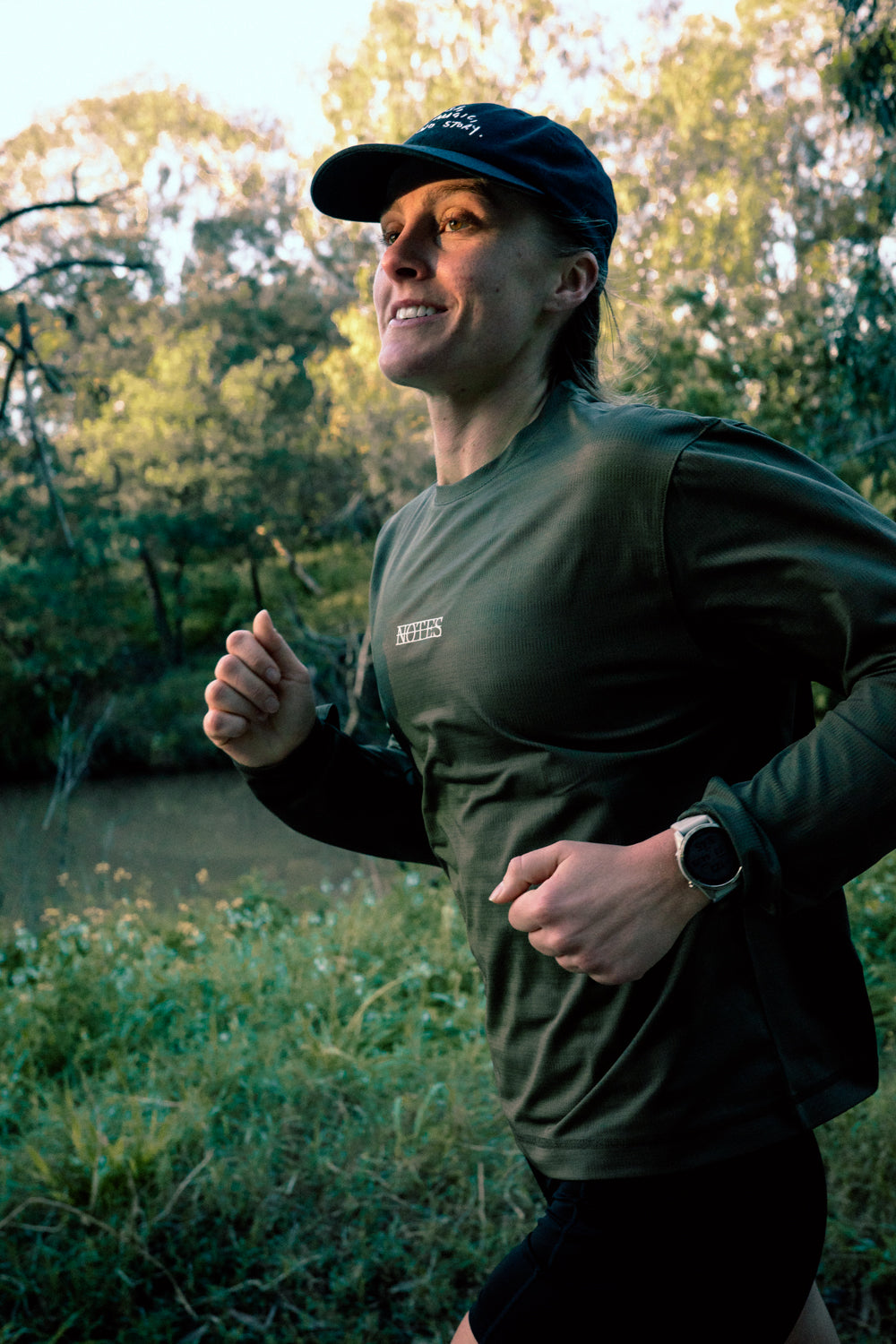 Runner in the No Risk No Magic No Story Cap in Washed Black and the Long Note LS in Olive on a riverside trail